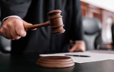 Ein Mann in schwarzer Robe hält einen Richter-Hammer in der Hand. Er ist durch einen Schreibtisch an dem er steht verdeckt und im oberen Bereich abgeschnitten. Im Hintergrund kann man die holzgetäfelte Rückwand eines Gerichtssaals leicht unscharf erkennen.