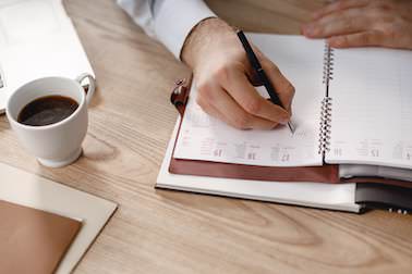 Ein Mann notiert etwas in einen in braunes Leder gebundenen Terminplaner, dabei ist nur seine Hand zu sehen. Der Planer liegt auf einem hellen Holztisch, auf dem eine fast volle Kaffeetasse steht, einige weitere Papiere sind angeschnitten zu erkennen.